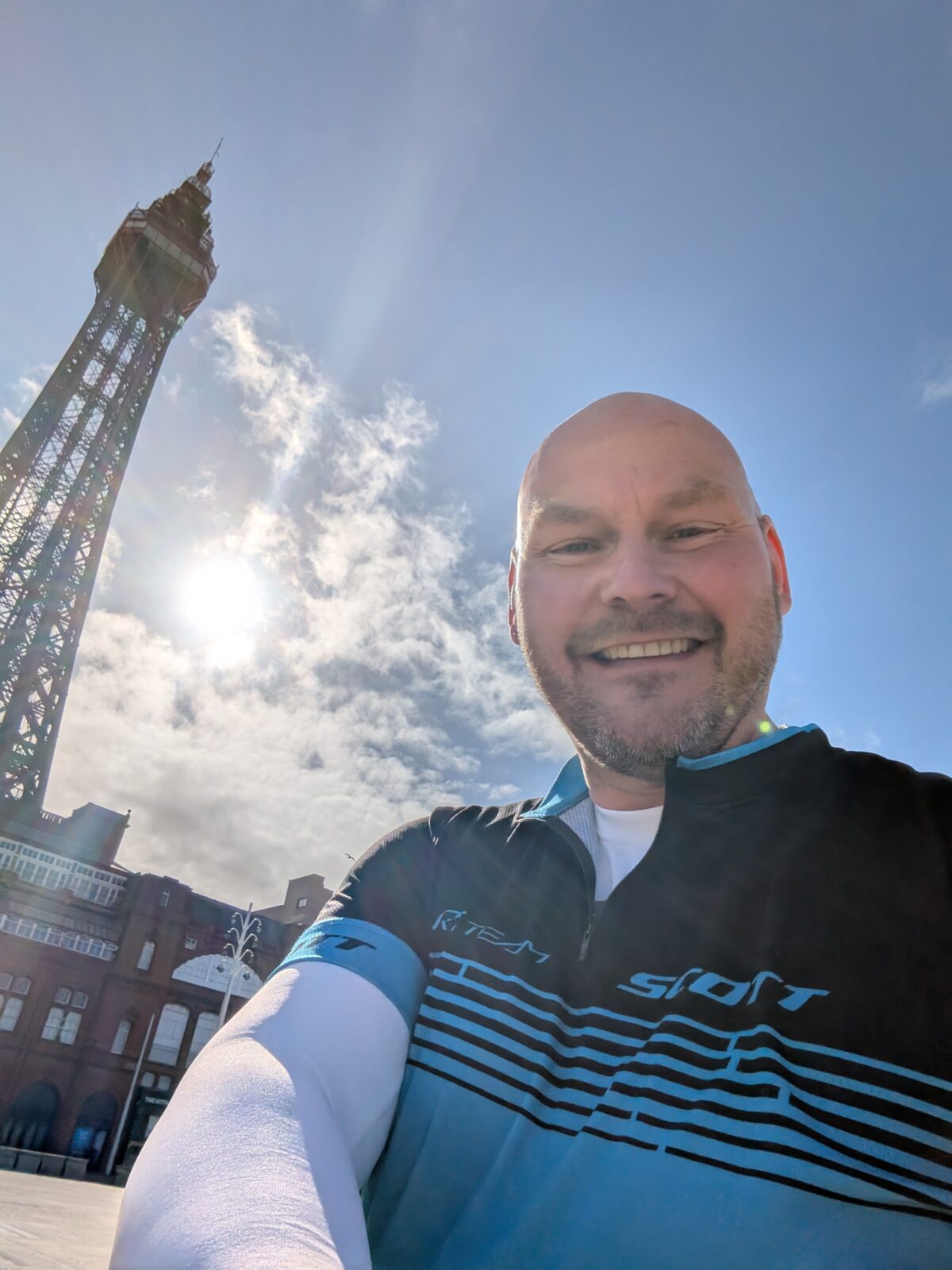 Smiling cyclist taking a selfie in front of Blackpool Tower at 9am, with the morning sun shining brightly through scattered clouds in a blue sky. The cyclist is wearing a blue and black jersey and appears cheerful at the start of the day's ride