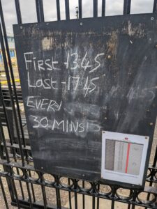 A weathered chalkboard sign at the Fleetwood ferry jetty displaying handwritten ferry times: "First - 13:45, Last - 17:45, Every 30 mins." A printed ferry timetable is also pinned to the bottom right corner of the board, all mounted on a black metal fence.
