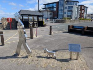 A polished metal sculpture of a man and his dog, titled "Matchstick Man and His Dog – LS Lowry," located on the Knott End promenade. The figures face towards the sea, set against a background of modern apartments and a small black shelter. The sculpture marks the spot where the artist LS Lowry sketched scenes of the ferry and coastline in the 1940s and 1950s, which inspired his painting "Jetty at Knott End."