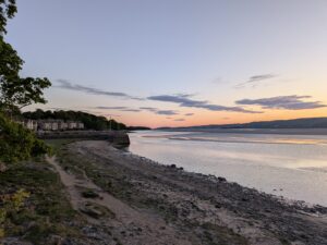 View of the shoreline at Arnside, Cumbria, at sunset. A row of houses lines the left-hand side along the coast, while calm water reflects the soft orange and pink hues of the sky. Low tide reveals a rocky, sandy beach in the foreground, with gentle hills visible in the distance across the estuary.