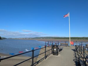 A Union Jack flag flutters on a tall flagpole at the end of a promenade in Arnside, overlooking the estuary. Red, white, and blue bunting is strung along black railings, and benches and bins line the viewing area. The sky is bright blue with scattered clouds, and low hills stretch across the horizon beyond the wide, sandy bay.
