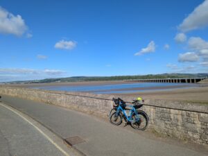 A blue touring bicycle leans against a stone wall on a seafront promenade in Arnside, Cumbria. Behind it, a long viaduct stretches across a wide expanse of sand and shallow water under a bright blue sky dotted with fluffy clouds. Hills and scattered houses are visible in the distance.