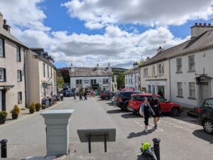 The village square in Cartmel, Cumbria, bustling with people under a partly cloudy sky. The Kings Arms pub faces the camera at the far end, with traditional whitewashed buildings and colourful bunting lining the street. Visitors walk and sit outside cafés and shops, including one named ‘Perfect English.’ Parked cars line both sides of the road.