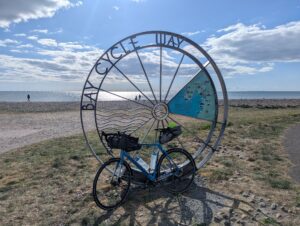 A touring bicycle leans against a large circular metal sculpture marking the Bay Cycle Way on Walney Island, Cumbria. The sculpture resembles a bicycle wheel and includes a map and wave design. Behind it, a shingle beach meets a shimmering sea under a partly cloudy sky, with distant wind turbines visible on the horizon.