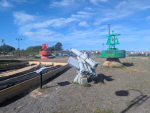 Outdoor maritime display at the Dock Museum in Barrow-in-Furness, featuring large red and green navigational buoys marked 'PRE' and 'EAST', along with a grey naval missile launcher on a gravel area under a blue sky. Raised beds and an information sign are visible in the foreground, with houses and lamp posts in the background.