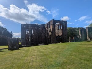 Ruins of Furness Abbey in the morning sunshine, with warm light casting shadows across the well-kept lawn. The large Gothic arches and stone walls of the historic structure stand partially intact, with some sections supported by scaffolding. Surrounded by trees and a bright blue sky with scattered clouds, the abbey exudes a serene, timeless atmosphere.
