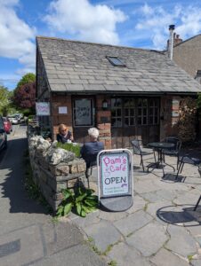 Pam’s Café in Kirkby-in-Furness, a small stone building with a slate roof and a cosy outdoor seating area. Two customers sit at a table beside a low stone wall, while a bright sign in front reads ‘Pam’s Café – OPEN – Muddy Boots & Dogs Welcome’. The café has rustic charm, with logs stacked neatly by the wall and a mix of sunshine and shade under a partly cloudy blue sky.