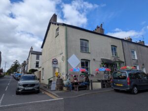 Street view of The Square Café in Broughton-in-Furness, Cumbria. The café occupies a pale cream corner building with green-trimmed windows and outdoor seating under colourful parasols. People sit at picnic tables enjoying food and drinks. Bunting decorates the front, and parked cars line the quiet street under a partly cloudy sky.