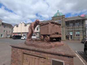 Bronze-coloured statue of a miner pushing an iron ore wagon, mounted on a brick plinth in a town square in Millom, Cumbria. Behind the statue is a building with a green-domed clock tower and scaffolding, with shops, cars, and overcast skies completing the scene. The statue commemorates the Hodbarrow Miner and the town’s heritage of mining and ironworks, as noted on a plaque below.