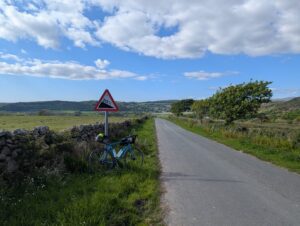 Quiet country road above Broad Oak in Cumbria, heading north through green fields and rolling hills under a blue sky with scattered clouds. A blue touring bicycle is propped against a stone wall beside a road sign warning of a 20% gradient. Trees line the roadside ahead, and the landscape stretches into wooded hills in the distance.