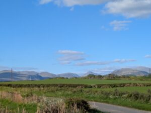 View of the Lake District mountains taken from the south, with a foreground of green fields, hedgerows, and a narrow country lane. The peaks rise in the distance under a clear blue sky with scattered clouds, with the distinctive shape of Scafell Pike and neighbouring fells visible. Electricity pylons dot the landscape, blending modern infrastructure with natural beauty.